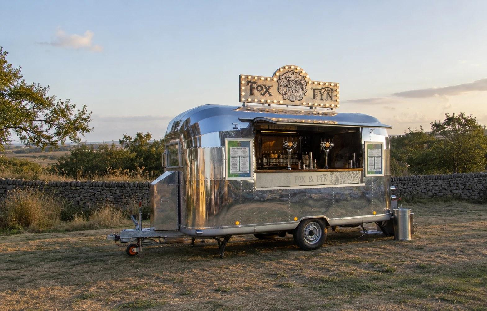 The Fox & Fyn Airstream in a meadow at sunset behind a drystone wall, its bulb-lit roof sign glowing and the bar set out along the open hatch