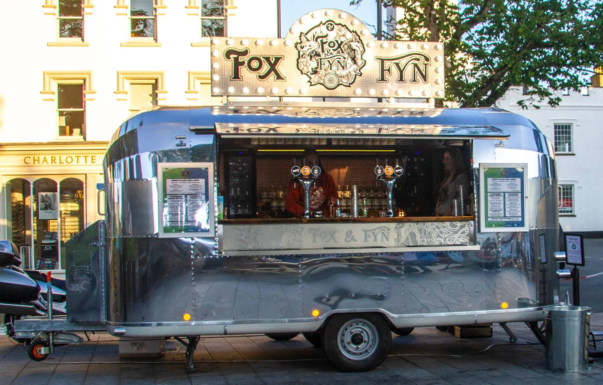 The Fox & Fyn Airstream at dusk in a town square, the lit sign on its roof glowing, the hatch folded out and two staff serving behind the counter